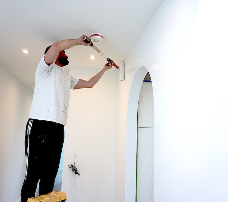 Interior painting by Kitsilano Painters. A professional painter on a ladder carefully painting the ceiling and walls of a modern white room, ensuring a smooth and flawless finish.